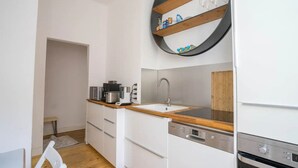 A compact kitchen with a sink, a stove, and a dishwasher. There is a wooden countertop and a white cabinet. A round shelf with bottles and glasses is mounted above the sink. - pestahome - Private Apartment Linden / Hanover, pestahome - Stadtwohnung in Linden (Hanover)