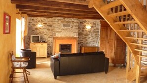 Interior rústico con pared de piedra, vigas de madera, chimenea, sofá, sillón de madera y televisor.