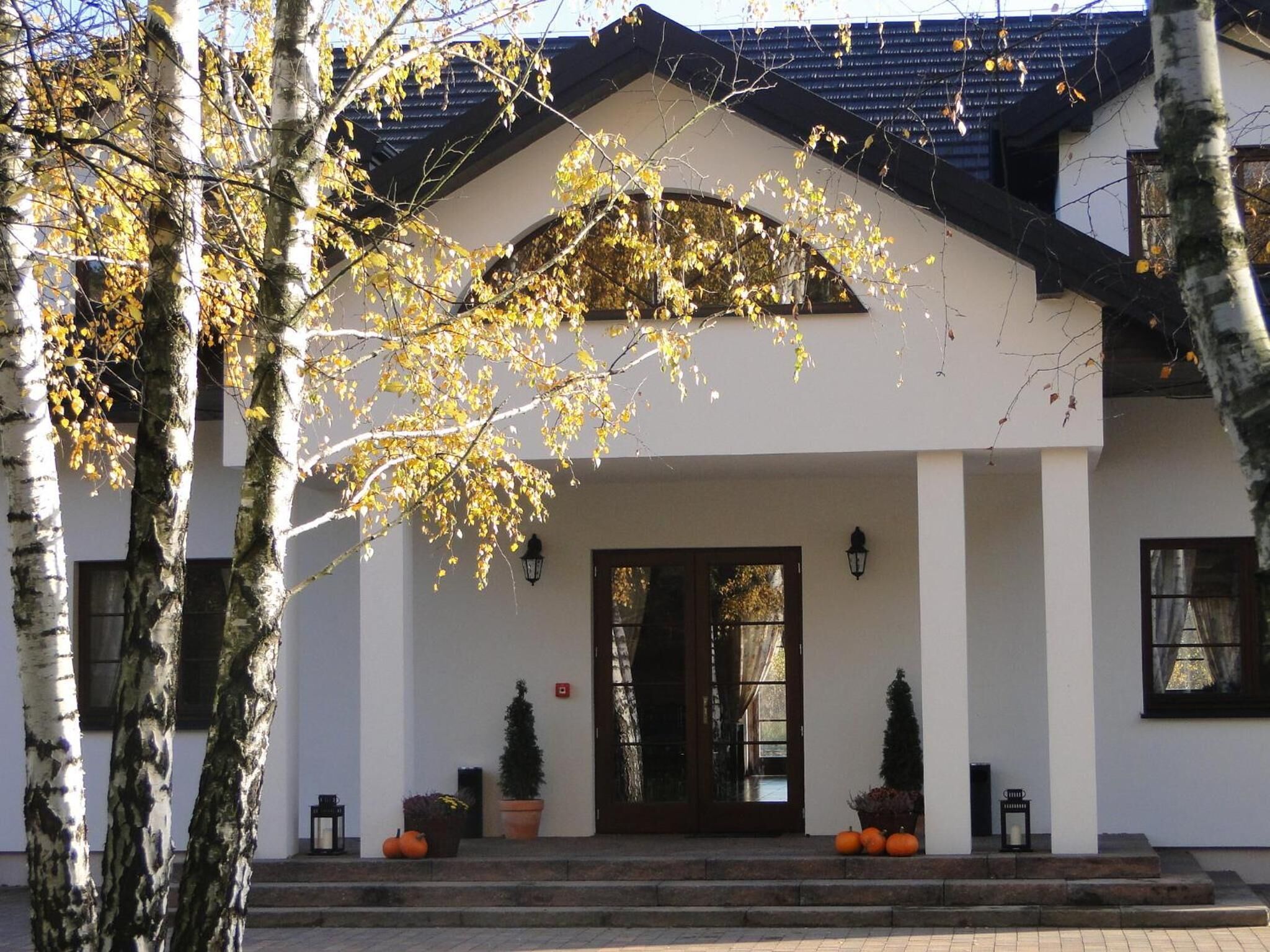 A building with a white facade, a dark roof, and a central entrance with a glass door. There are two potted plants and a small tree on the steps.