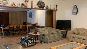 Ein Wohnzimmer mit Fernseher, einer Couch, einem Sessel und einem Essbereich mit Stühlen und einem Tisch.