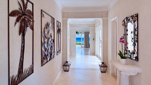 A hallway with decorative metal wall art, a white stone bench, and a view of the ocean through an open doorway. - Stylish Beachfront Villa with Plunge Pool - Still Fathoms (4 bed) (Reeds Bay)