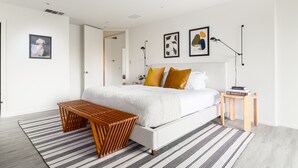 Ein modernes Schlafzimmer mit einem weißen Bett, einer Holzbank, einem Streifen Teppich und einem kleinen Holznachttisch mit Büchern.