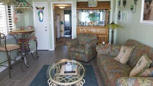 Ein Wohnzimmer mit einer Couch, einem Couchtisch, einem Esstisch mit Stühlen, einem Deckenventilator und einer Küche im Hintergrund.