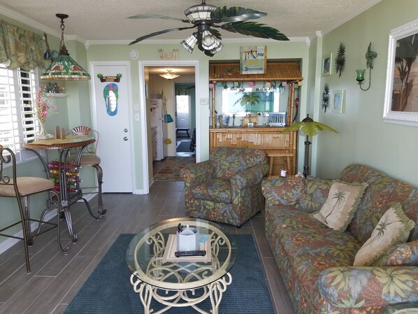 A living room with a sofa, a coffee table, a dining table with chairs, a ceiling fan, and a kitchen area in the background. - Dave's Vacation Rentals - Palms resort (Myrtle Beach)