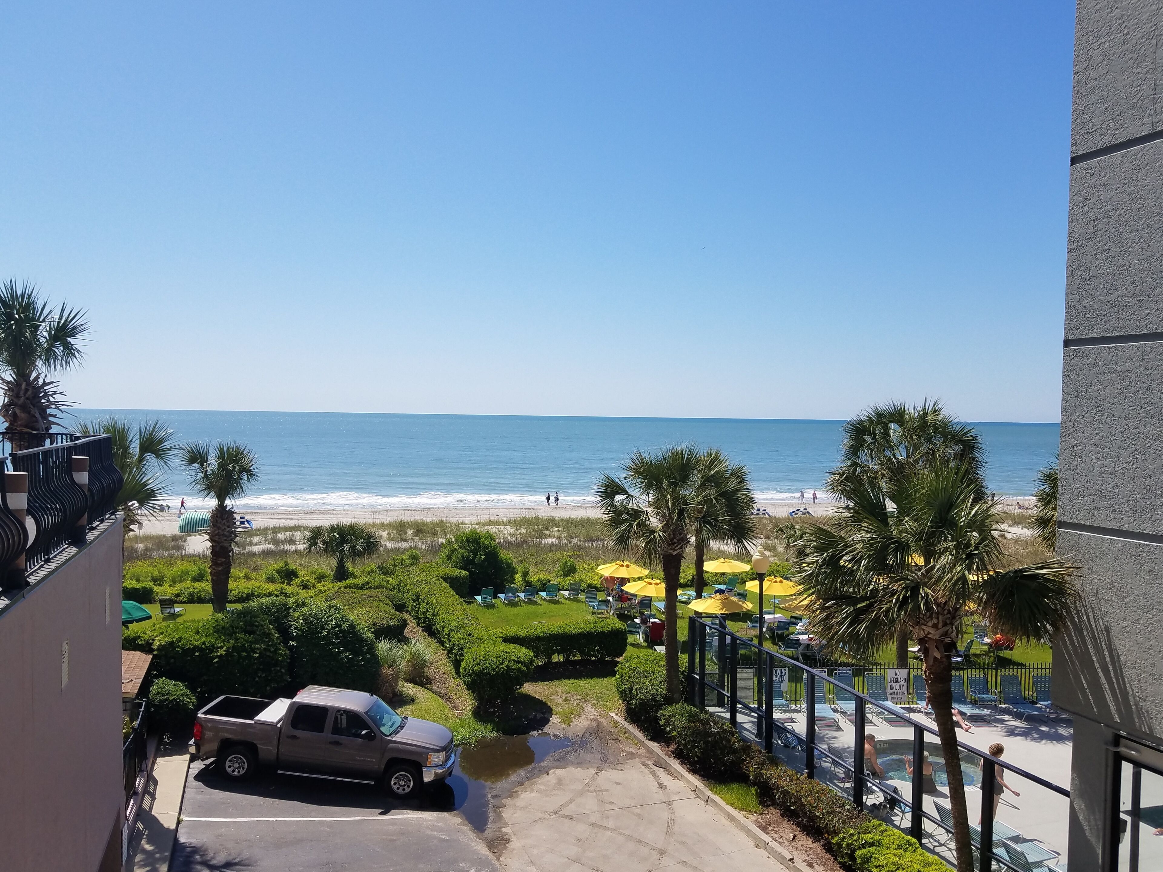 Blick auf einen Strand von einem Balkon aus, mit einem abgestellten Lastwagen, einem Poolbereich mit Sonnenschirmen und einem strahlend blauen Himmel.