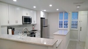 A modern kitchen with white cabinets, stainless steel appliances, a marble countertop, and a window with blinds. - Beautiful Marco Island Renovated Condo, 2 Br 2 Ba, 1704 T4,  Beach & Ocean Views (Marco Island)