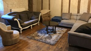 A cozy living room with a stone wall, wooden beams, a grey sofa, a patterned armchair, a small round table, and a woven chair.