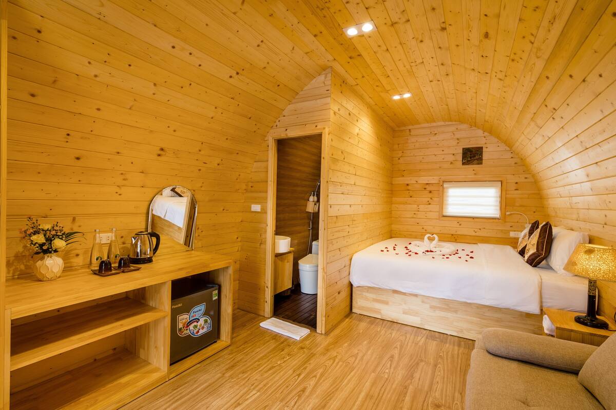 A wooden cabin room with a bed, a small seating area, a wooden shelf with items, and a window.