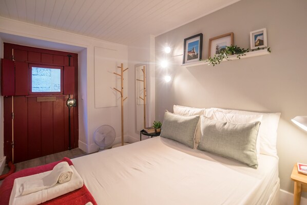 A neatly arranged bedroom with a bed, a red door, a small bedside table with a plant, and a wall-mounted shelf with pictures.