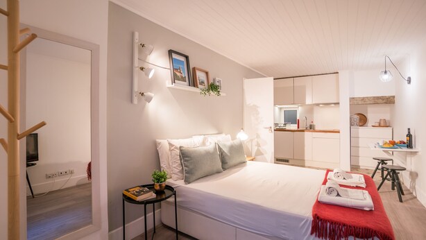 A modern bedroom with a large bed, bedside tables, a small side table with a plant, a red throw blanket, and a view into the adjacent kitchen. - Rose 7 Studio Apartment (Lisboa)
