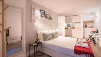 Quarto moderno com uma cama grande, mesas de cabeceira, uma mesinha lateral com uma planta, um cobertor vermelho e vista para a cozinha adjacente.