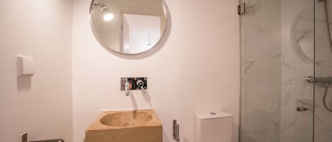 A bathroom with a round mirror, a beige sink, a toilet, and a glass shower enclosure.