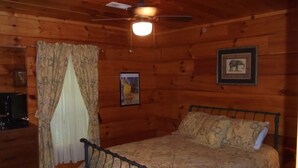 A wooden cabin room with a bed, a ceiling fan, a framed picture, and a television on a wooden dresser.