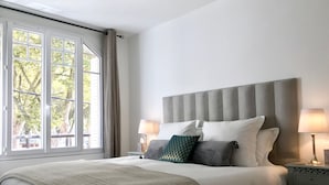 A neatly made bed with a grey headboard, two bedside lamps, and a view of trees through large windows. - Luxurious Vacation Rental 10 mins walk from Epernay town centre (Épernay)