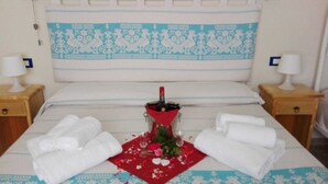 A neatly made bed with a decorative headboard, two bedside tables with lamps, and a bottle of champagne on a red napkin. - Meravisola (Castiadas)