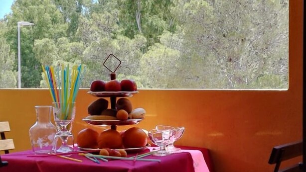 A table with a pink tablecloth, a fruit platter, and a glass of water on a balcony with a view of trees. - Meravisola (Castiadas)