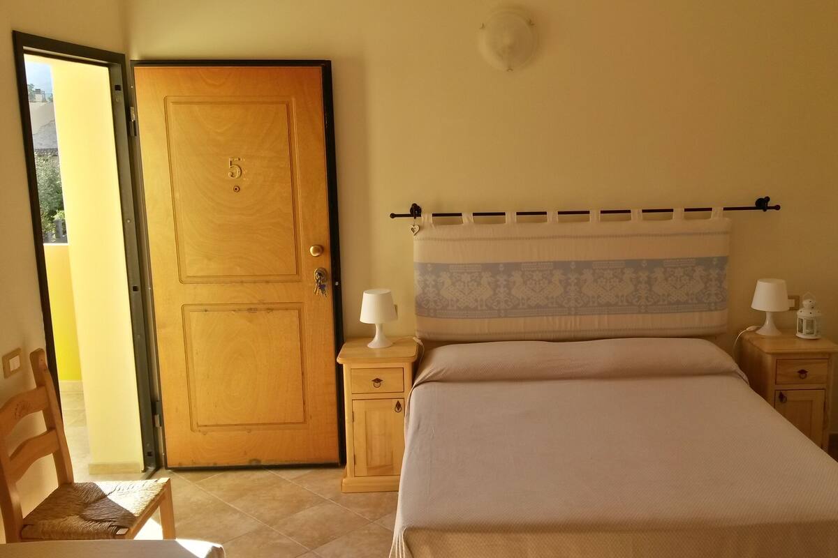A bedroom with a wooden door, a bed with a headboard, two bedside tables with lamps, and a chair.