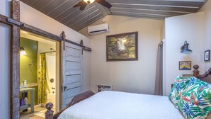 Un dormitorio con una cama, una puerta de madera, un equipo de aire acondicionado, un ventilador de techo, un cuadro en la pared y una lámpara de noche.
