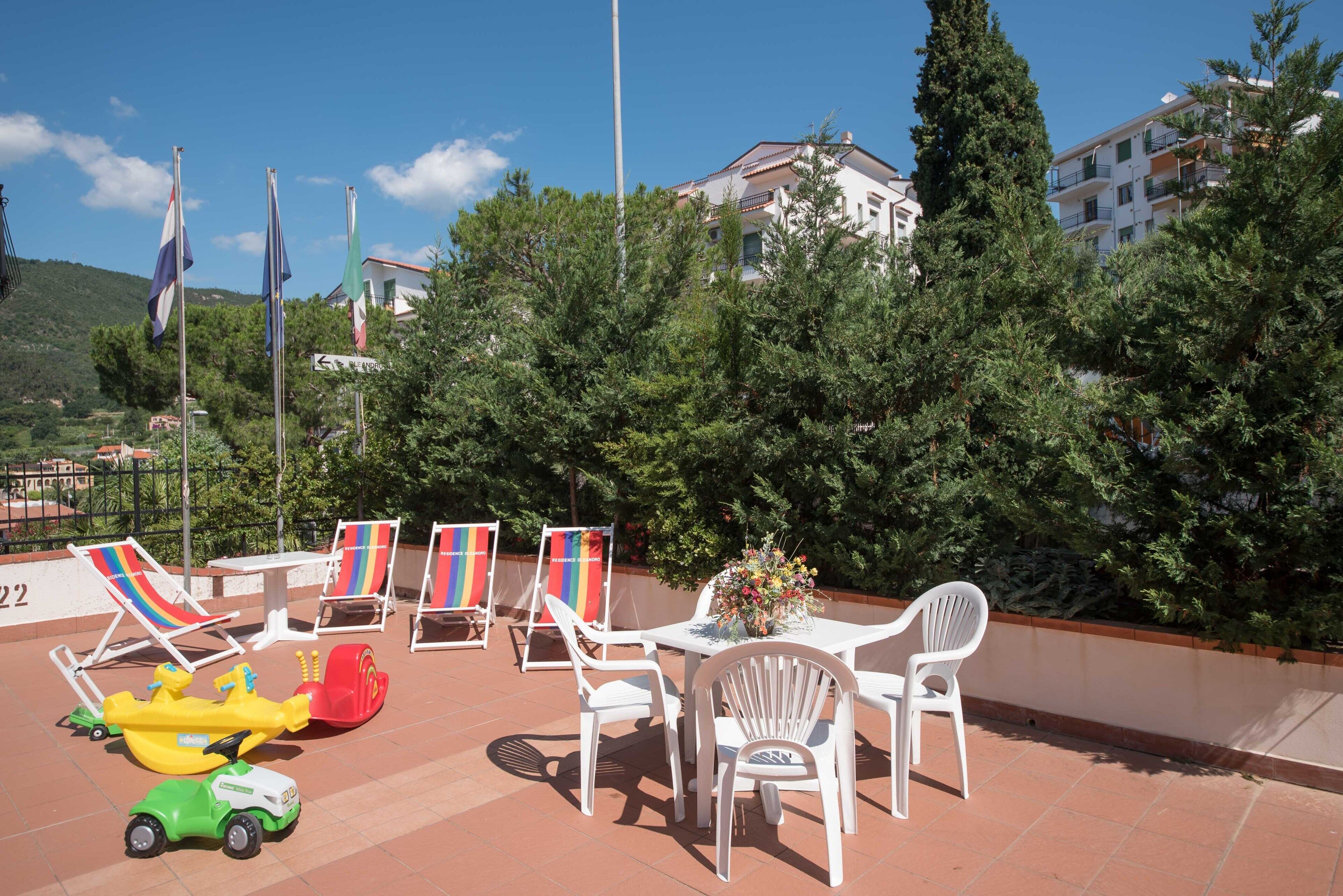 Un espacio al aire libre con sillas y mesas blancas, una zona de juegos infantil y banderas.