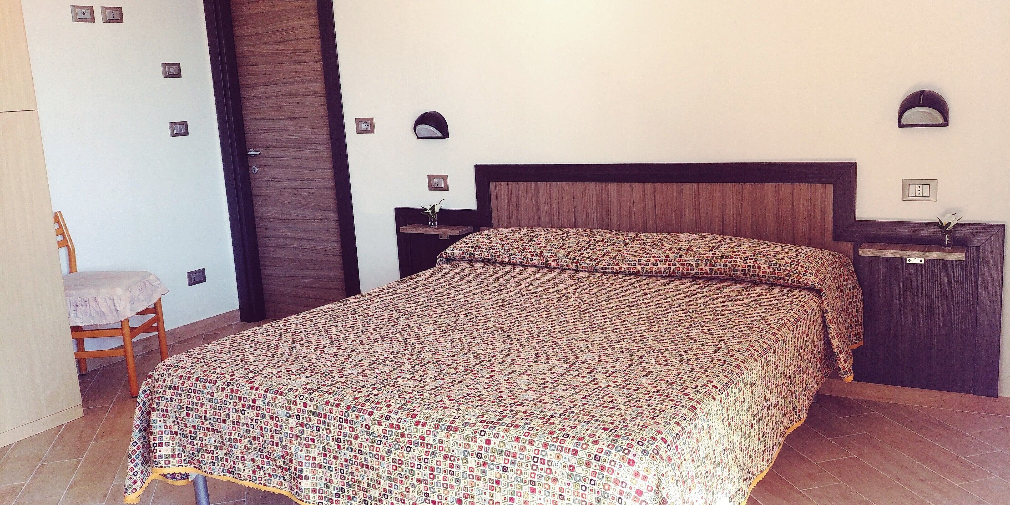 A neatly made bed with a patterned bedspread, a wooden nightstand with a lamp, and a chair with a cushion.