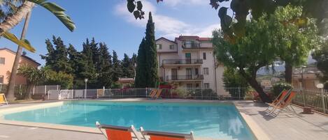 A swimming pool with lounge chairs, surrounded by a fence, with buildings and trees in the background.