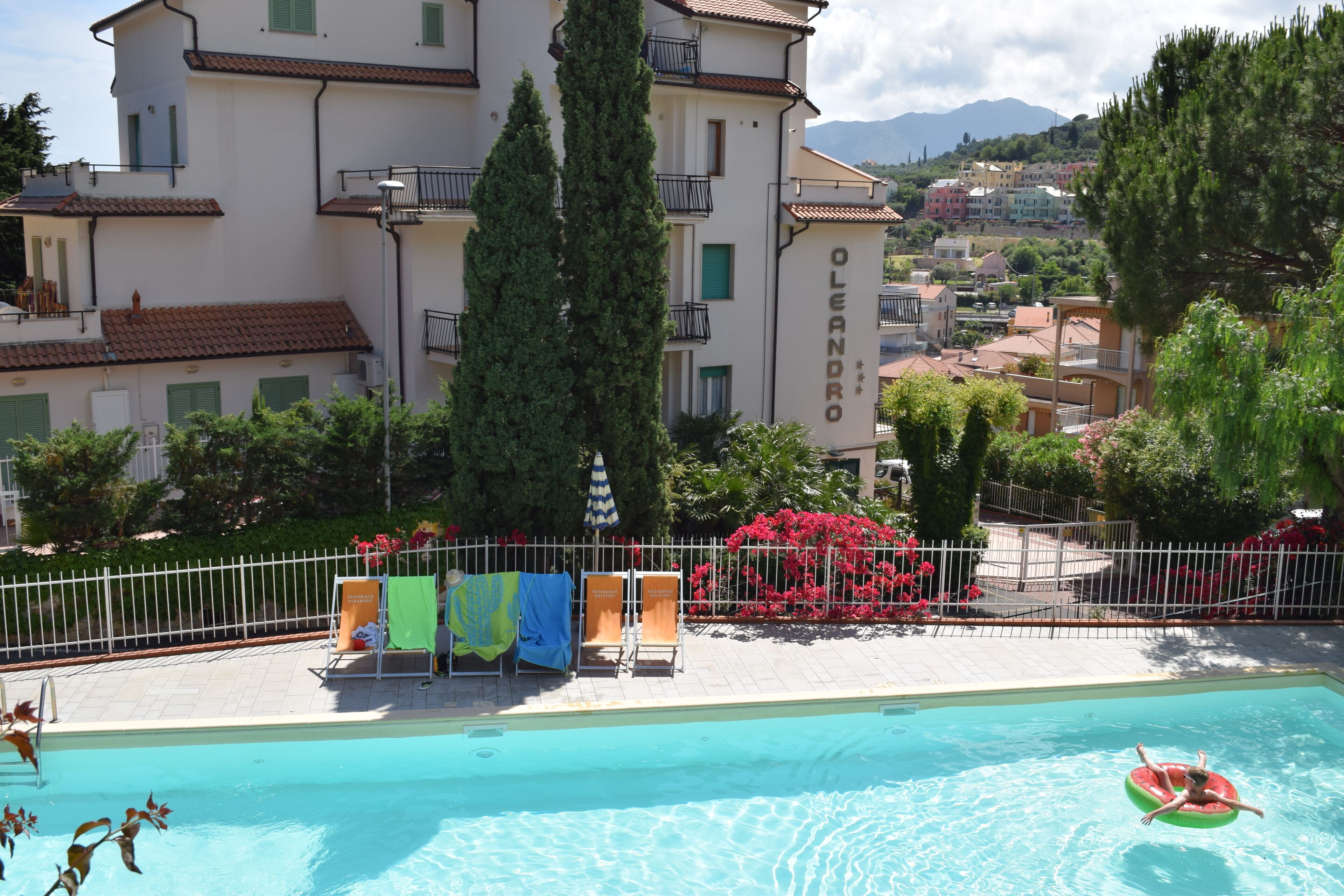 A swimming pool with lounge chairs and a fence, surrounded by greenery and buildings.