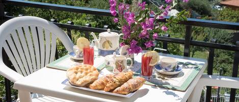 A balcony table with a white chair, a tray of pastries, a teapot, cups, and a vase of flowers.