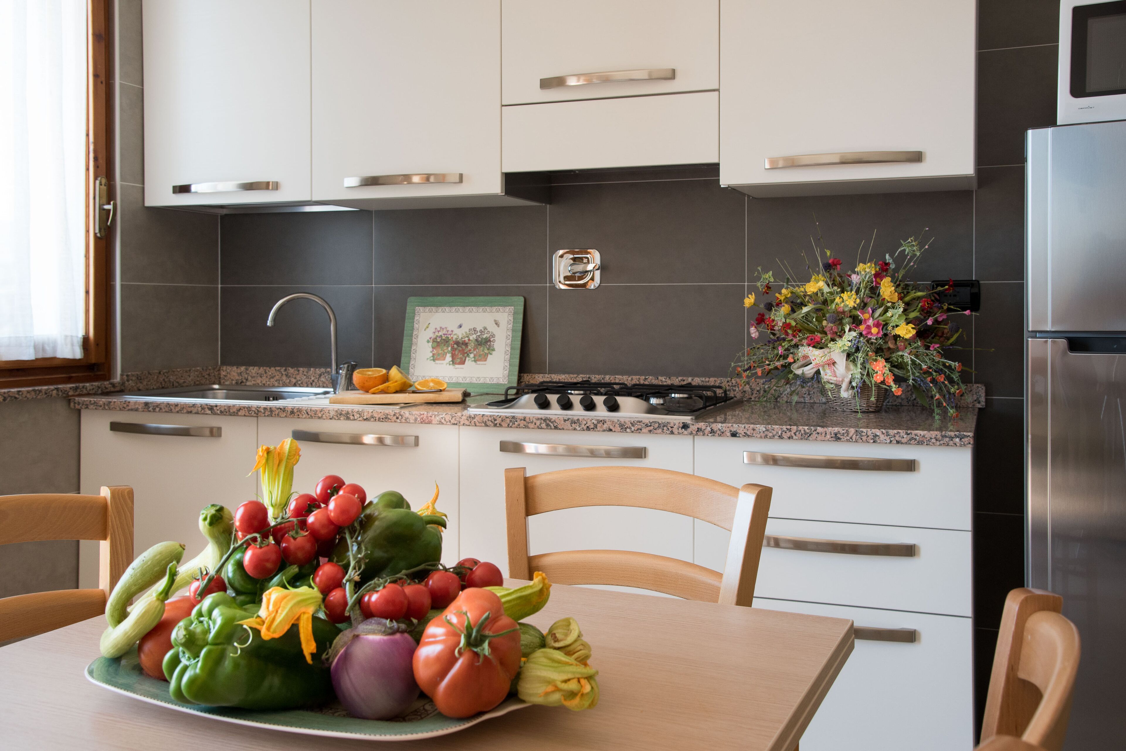 Una cocina con armarios blancos, un refrigerador de acero inoxidable y una mesa de comedor con un plato de verduras.