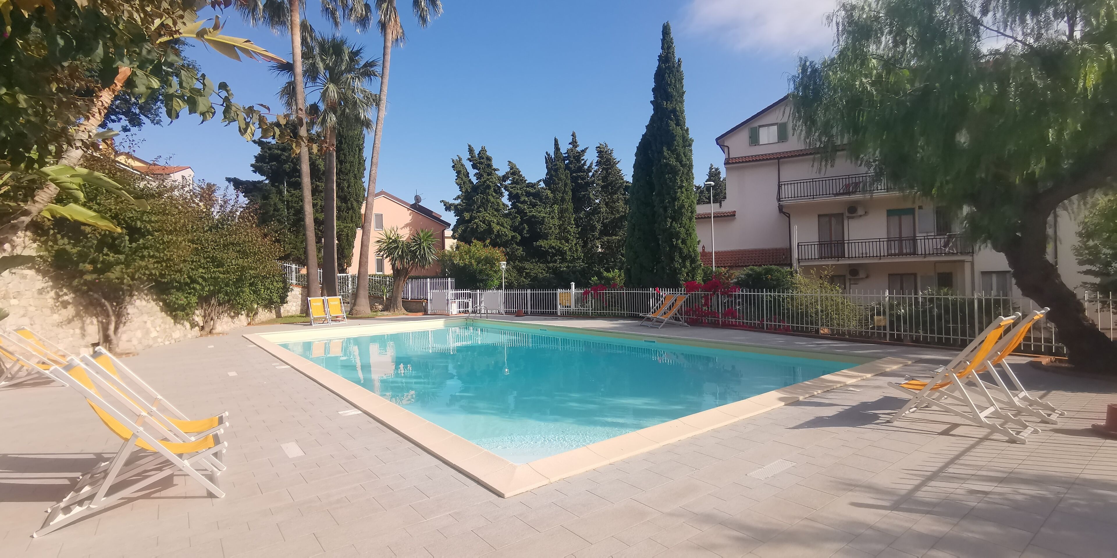 A rectangular pool with a tiled deck, surrounded by a metal fence and trees.