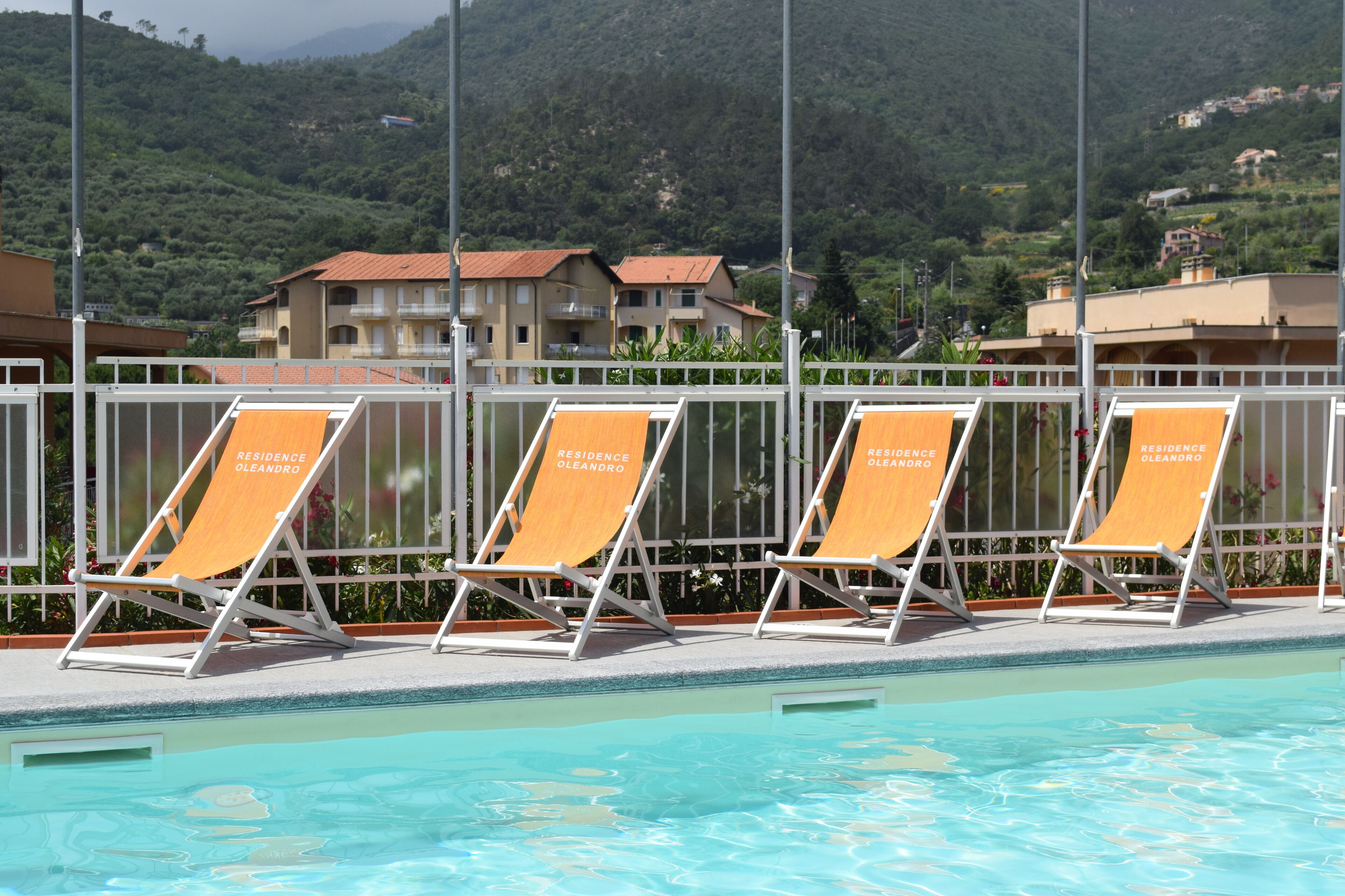 Un espace aménagé au bord de la piscine, avec des chaises longues portant l’inscription « Résidence Oleandro », une vue dégagée sur un paysage montagneux et une piscine dont les couloirs de nage sont clairement délimités.