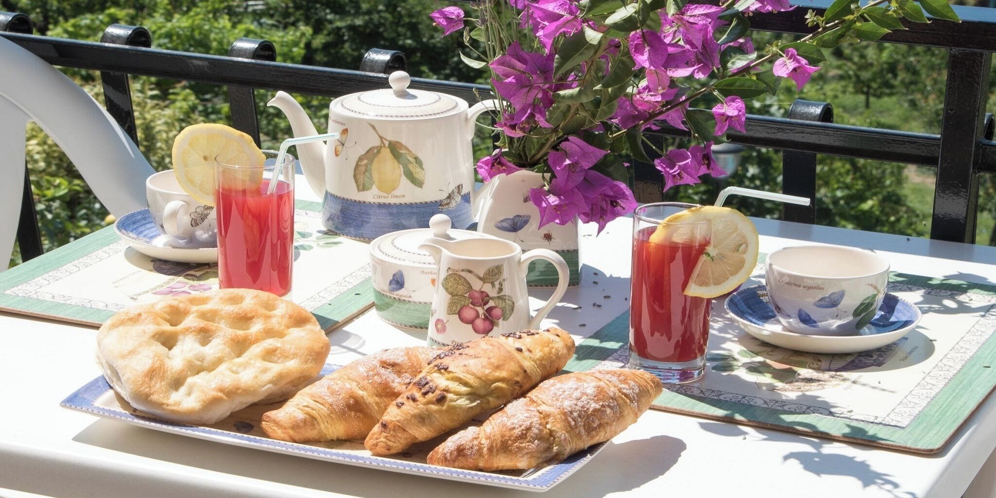 A table set with pastries, tea, and juice on a balcony with a view of greenery.