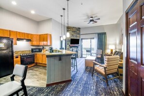 A modern kitchen with wooden cabinets, a granite countertop, and a built-in island. There is a dining area with a bar stool and a chair. The room has a blue carpet and a ceiling fan.