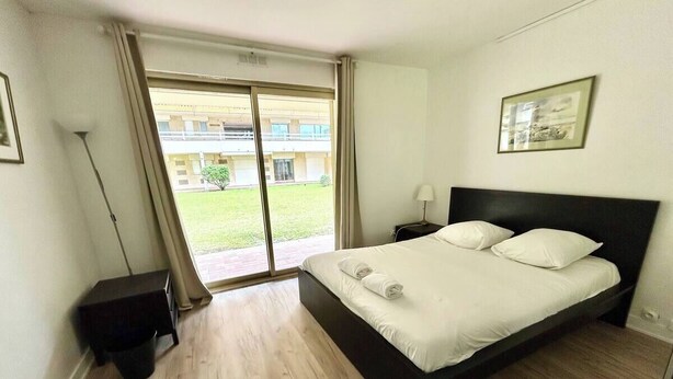 Een moderne hotelkamer met een groot bed, een klein nachtkastje, een lamp en uitzicht op een gebouw en groen via een schuifdeur.