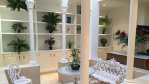 A well-lit indoor space with white columns, a round table, and a bench. Shelves with potted plants are visible in the background.