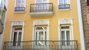 A yellow building with a wooden door, white-framed windows, and a balcony with metal railings.