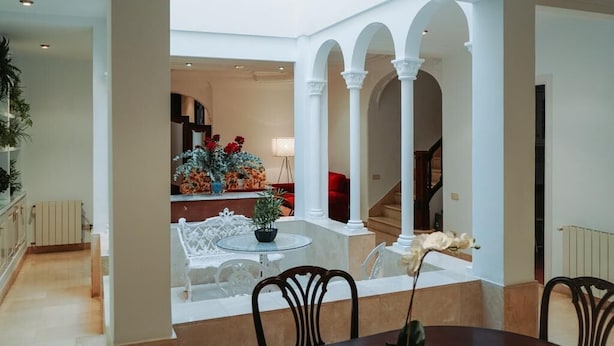 A spacious indoor area with a dining table, chairs, and a potted plant. There are white columns and arches, and a staircase is visible in the background.