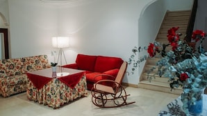 A living room with a red sofa, a rocking chair, a floral-patterned sofa, a small table with a lamp, and a staircase with red flowers.
