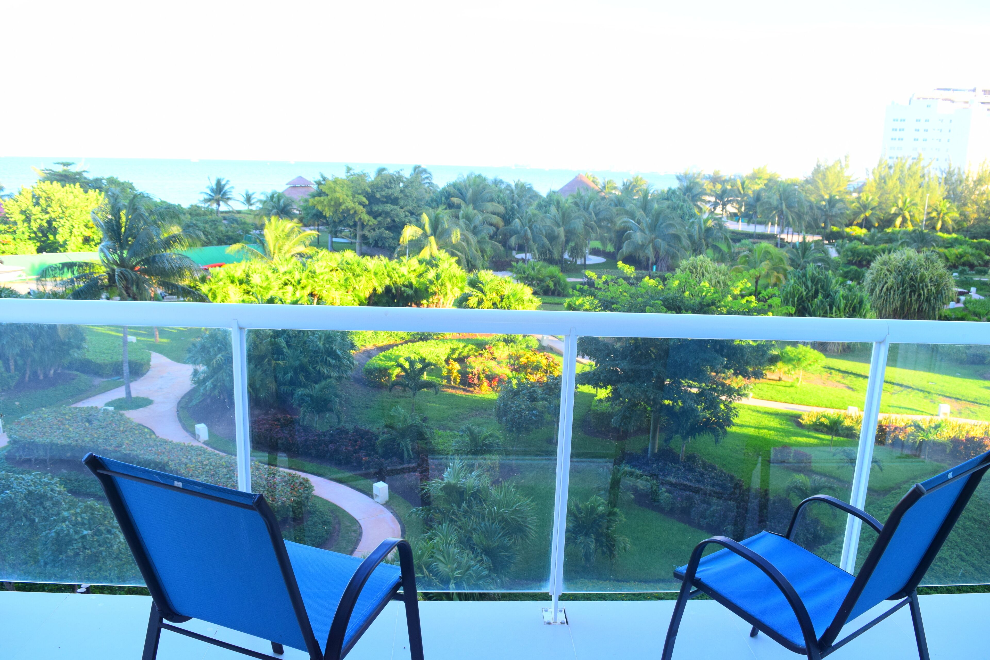 A balcony with two blue chairs and a glass railing overlooking a landscaped garden with palm trees and a view of a building in the distance.