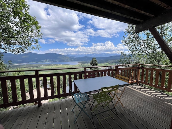 A wooden deck with a table and chairs, overlooking a scenic landscape with mountains and a lake.