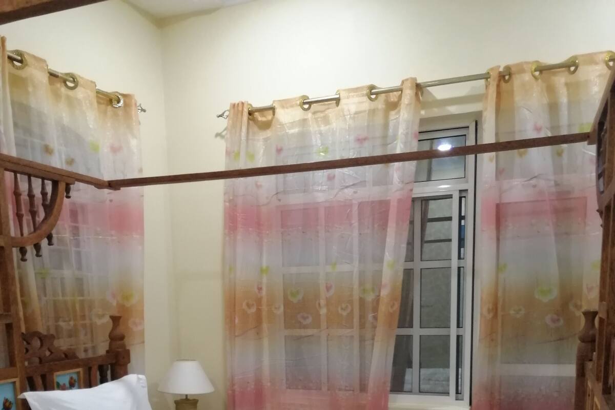 Un lit bien fait, agrémenté d’une serviette blanche décorative, d’une tête de lit en bois et d’une fenêtre ornée de rideaux à motifs floraux.