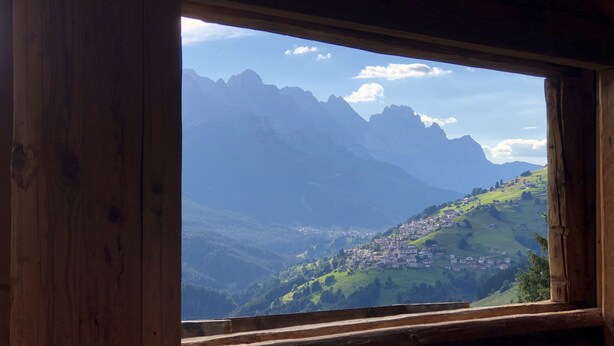 A wooden cabin with a large window offering a view of a mountainous landscape and a small village below.