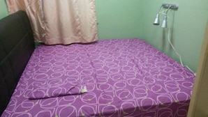 Una habitación pequeña con cama individual, colcha estampada color morado, cabecera, enchufe y cortina.