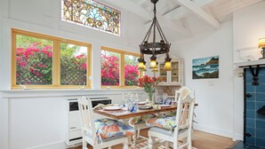 A dining area with a wooden table, white chairs, and a stained glass window above. - Seashore Retreat - Perfect Family Cottage by the Beach (Montecito)