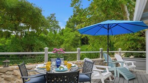 A deck with a table set for dining, surrounded by Adirondack chairs, and a blue umbrella providing shade. - Seashore Retreat - Perfect Family Cottage by the Beach (Montecito)