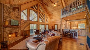 A spacious living area with a stone fireplace, leather sofa, and a large window overlooking a forest.