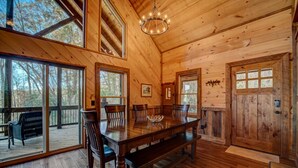 A wooden interior with a dining table and chairs, large windows, and a chandelier.