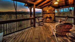 A wooden deck with a stone fireplace, wooden rocking chairs, and a view of the surrounding landscape.