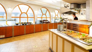 Montaje tipo bufé con varias estaciones de comida, un chef preparando alimentos y amplias ventanas arqueadas que dejan entrar luz natural.