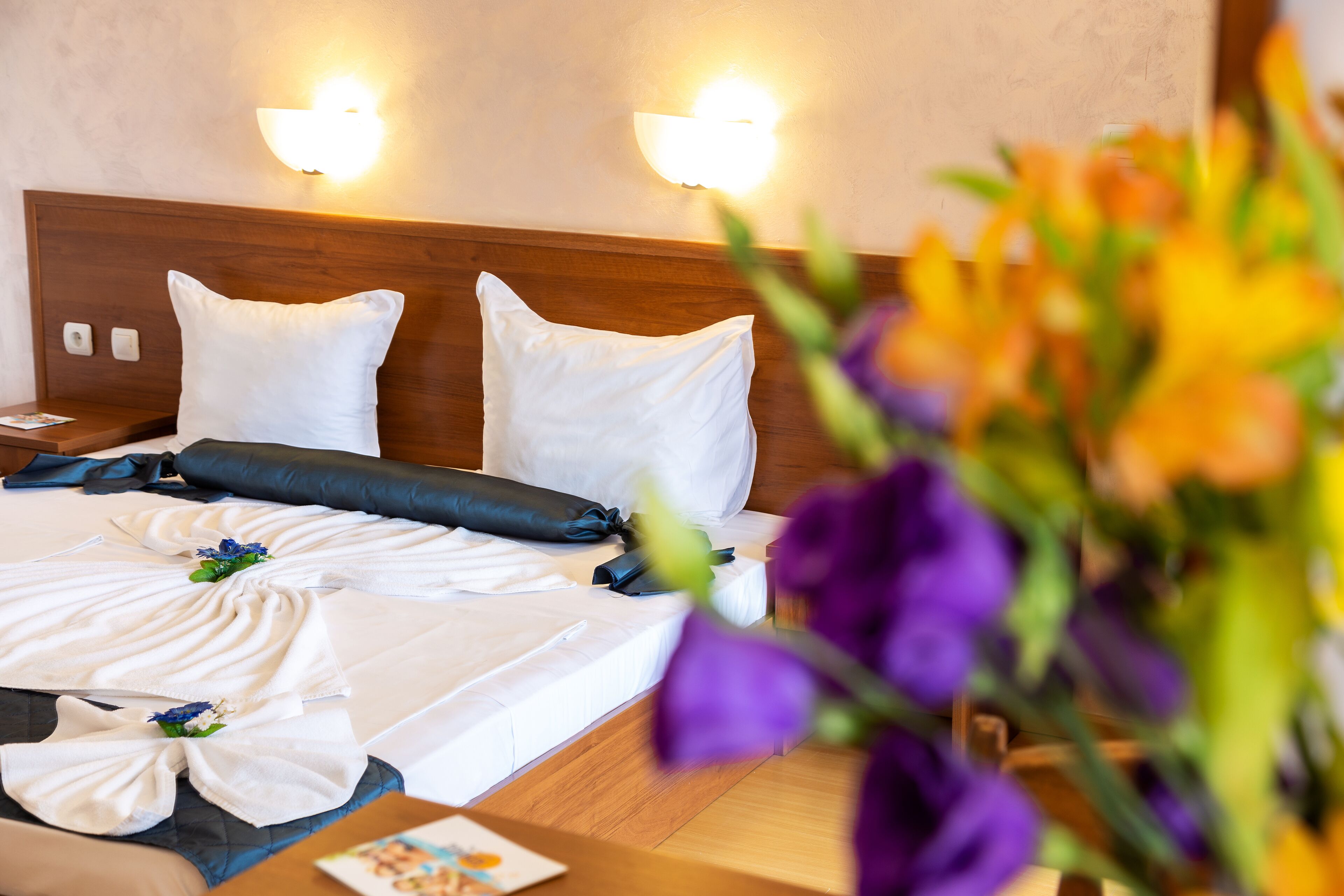 Een net opgemaakt bed met witte lakens en een donkerblauwe kussen, een houten hoofdbord en twee wandlampen.