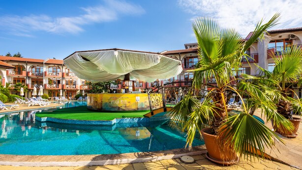 Piscina con sillas de descanso y bar junto a la piscina, rodeada de palmeras y un edificio de hotel.
